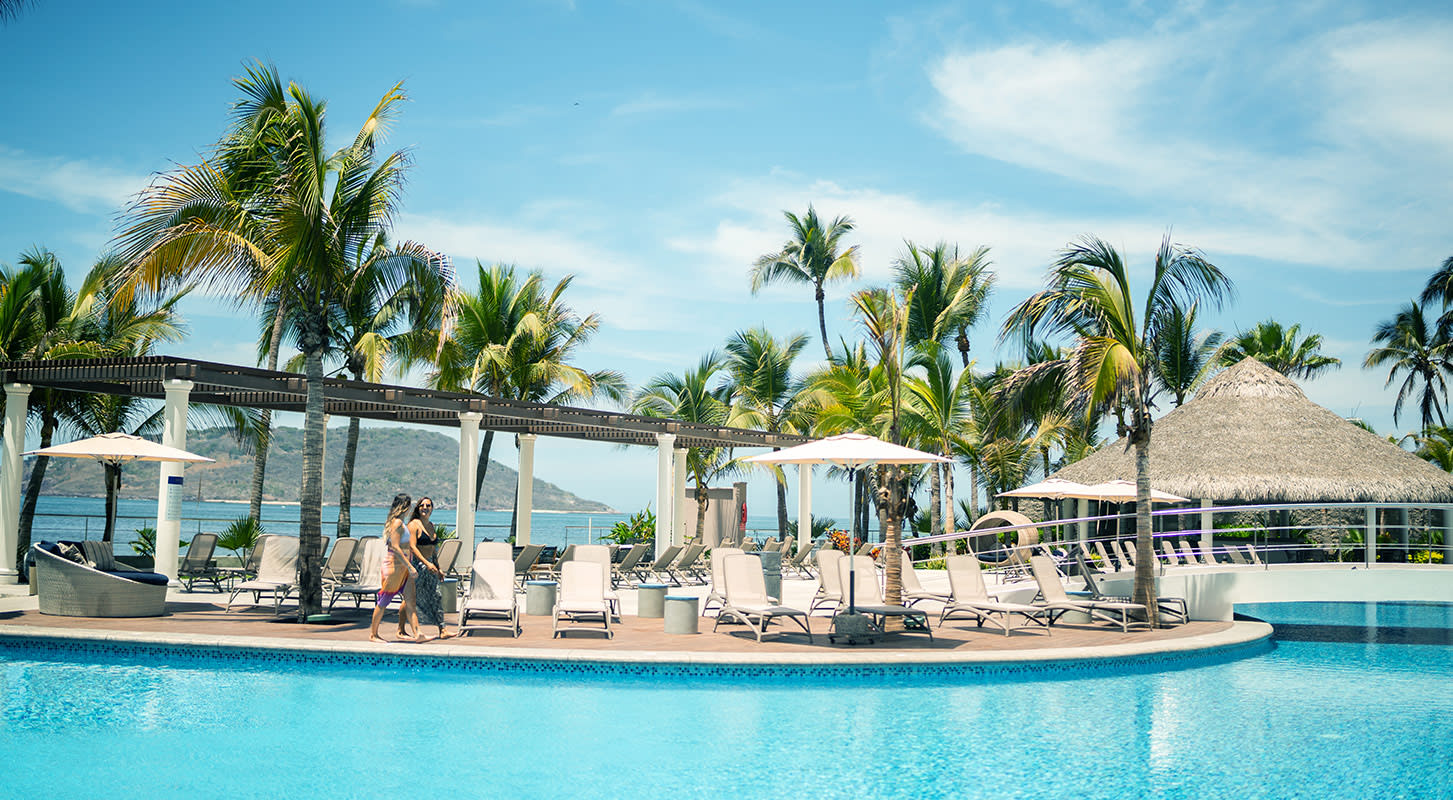 A serene pool area with lounge chairs and umbrellas, situated near a sandy beach under a clear blue sky.